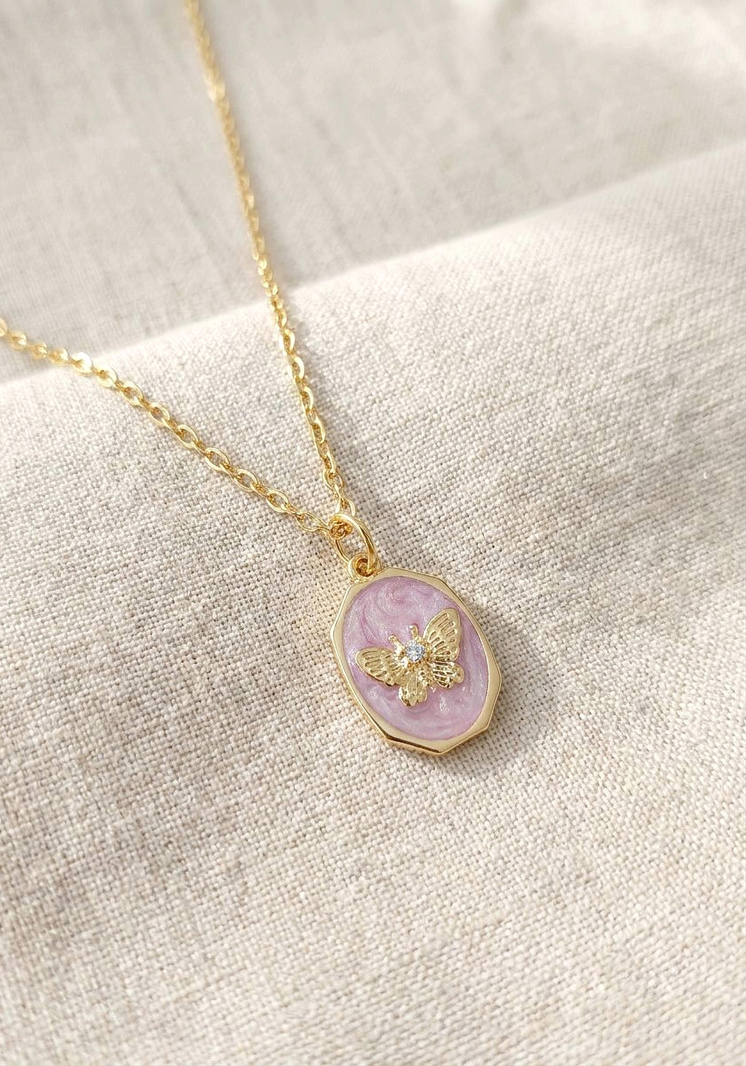 Collier papillon m&eacute;daillon rose et dor&eacute; pos&eacute; d&eacute;licatement sur un tissu en lin